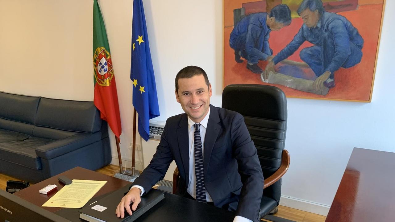 Ein junger Mann sitzt in die Kamera strahlend in seinem Büro am Schreibtisch, hinter ihm die Fahne Europas und Portugals. Ein junger Mann sitzt in die Kamera strahlend in seinem Büro am Schreibtisch, hinter ihm die Fahne Europas und Portugals.
