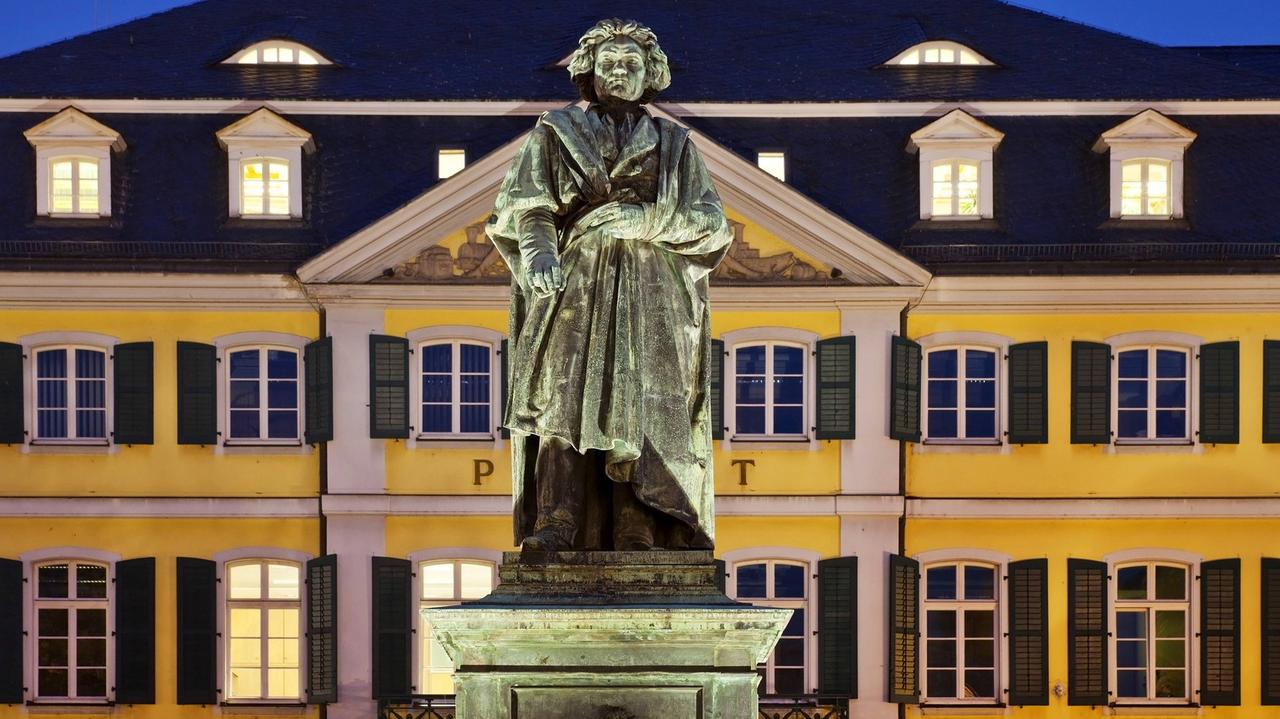 Im Vordergrund das Beethoven-Denkmal, im Hintergrund das Hauptpostamt Bonns. Das Denkmal besteht aus einer überlebensgroßen Darstellung Beethovens auf einem Sockel, das hier am Abend angestrahlt wird und sich daher deutlich von dem gelb gestrichenen mehrstöckigen Gebäude im Hintergrund abhebt.