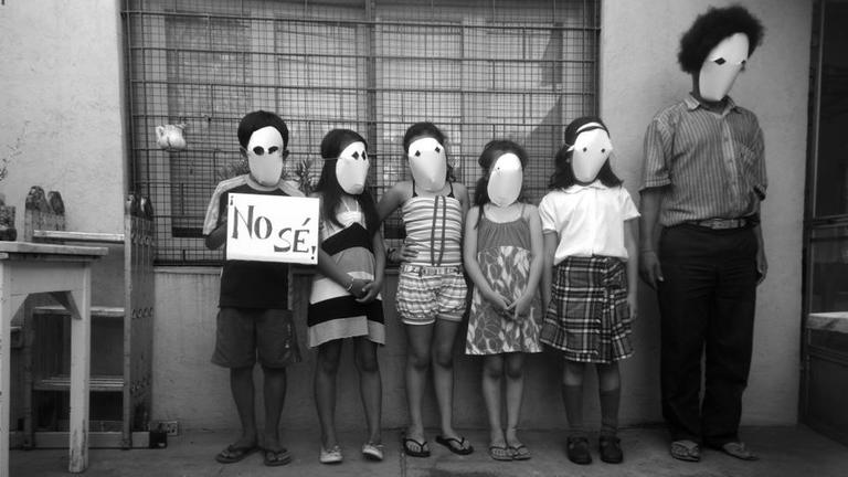 Schwarzweißfoto von Paulo Nazareth. Darauf zu sehen: fünf Kinder und ein erwachsender Mann in einer Reihe vor einer Häuserwand. Ihre Gesicht sind hinter weißen Masken verborgen. Ein Junge hält ein Schild mit der Aufschrift "No sé!".