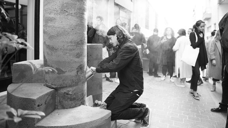 Eine Schwarz-Weiß Fotografie zeigt den Künstler Oscar Tuazon knieend bei der Arbeit an der Steinskulptur "Building Fire"