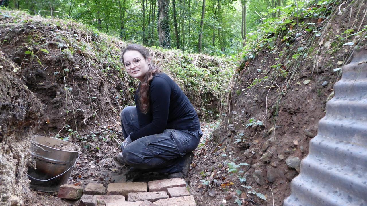 Eine Frau mit Arbeitskleidung sitzt in einem Graben und lächelt in die Kamera. Eine Frau mit Arbeitskleidung sitzt in einem Graben und lächelt in die Kamera.