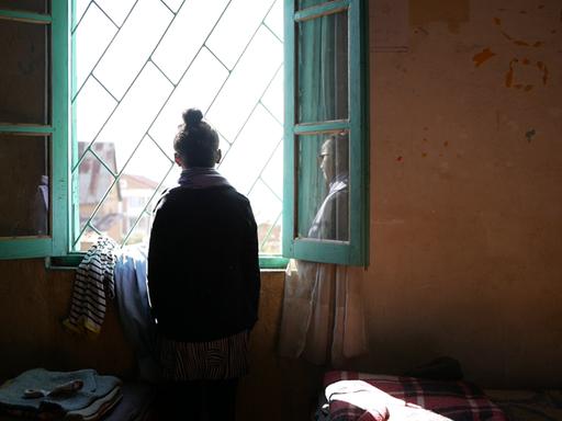 Melancholische Rückaufnahme eines jungen Mädchen, das aus einem Fenster nach draußen schaut.