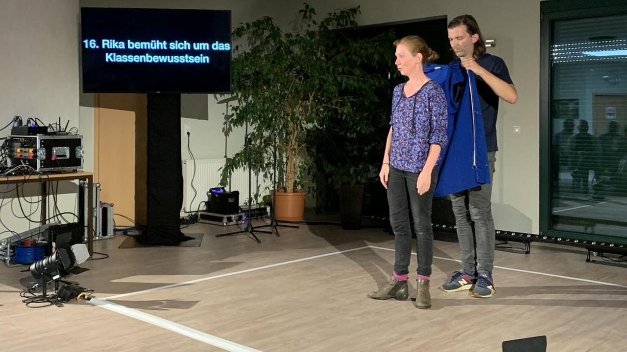 Rika Weniger steht in einem Raum. Auf dem Boden ist mit Klebestreifen ein großes Quadrat abgeklebt. Darin steht sie und ihr Bühnenpartner hilft ihr in eine blaue Jacke. Rika Weniger steht in einem Raum. Auf dem Boden ist mit Klebestreifen ein großes Quadrat abgeklebt. Darin steht sie und ihr Bühnenpartner hilft ihr in eine blaue Jacke.