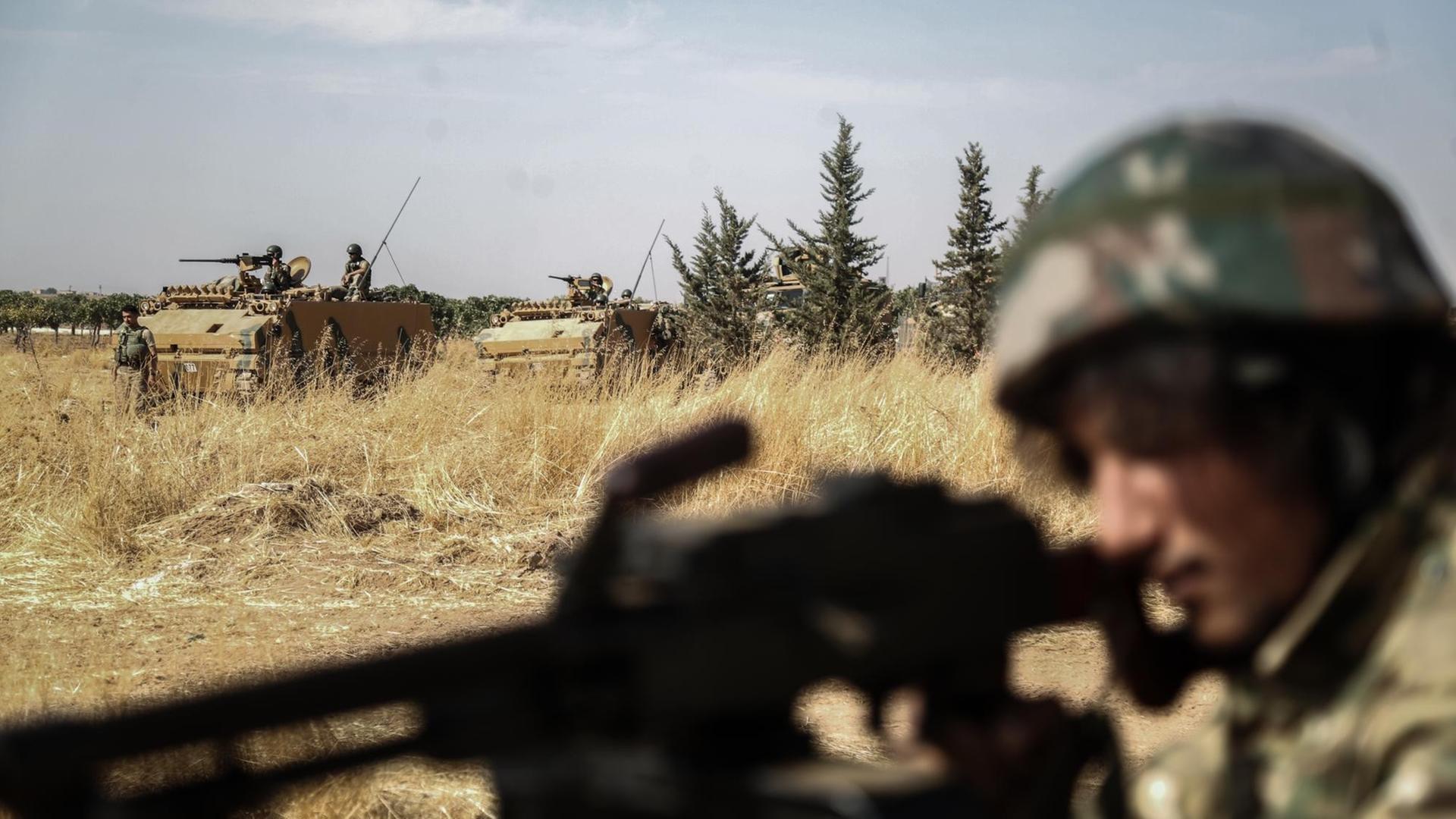 Ein von der Türkei unterstützte Rebell der Syrischen Nationalarmee richtet seine Waffe aus, während im Hintergrund türkische Panzer vorbeifahren Ein von der Türkei unterstützte Rebell der Syrischen Nationalarmee richtet seine Waffe aus, während im Hintergrund türkische Panzer vorbeifahren