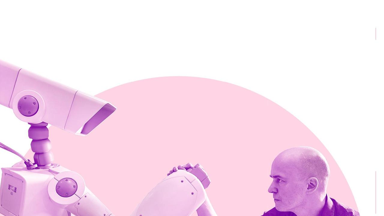 Ein Roboter und ein Mann mit kurzen Haaren sitzen sich an einem Tisch gegenüber und tragen einen Wettkampf im Armdrücken aus.