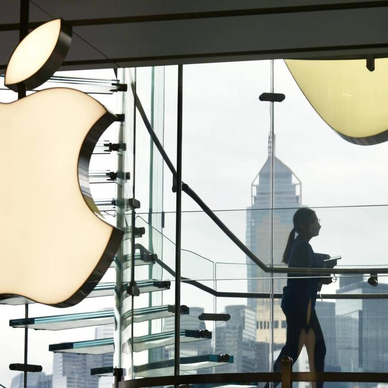 Eine Frau läuft in einem gläsernen treppenhaus an einem leuchtenden Apple-Logo vorbei. 