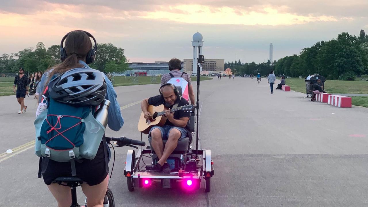 Eine Fahrradfahrerin mit Kopfhörern auf dem Tempelhofer Feld, vor ihr fährt ein Radfahrer mit Anhänger, auf dem ein Musiker Gitarre spielt. Eine Fahrradfahrerin mit Kopfhörern auf dem Tempelhofer Feld, vor ihr fährt ein Radfahrer mit Anhänger, auf dem ein Musiker Gitarre spielt.