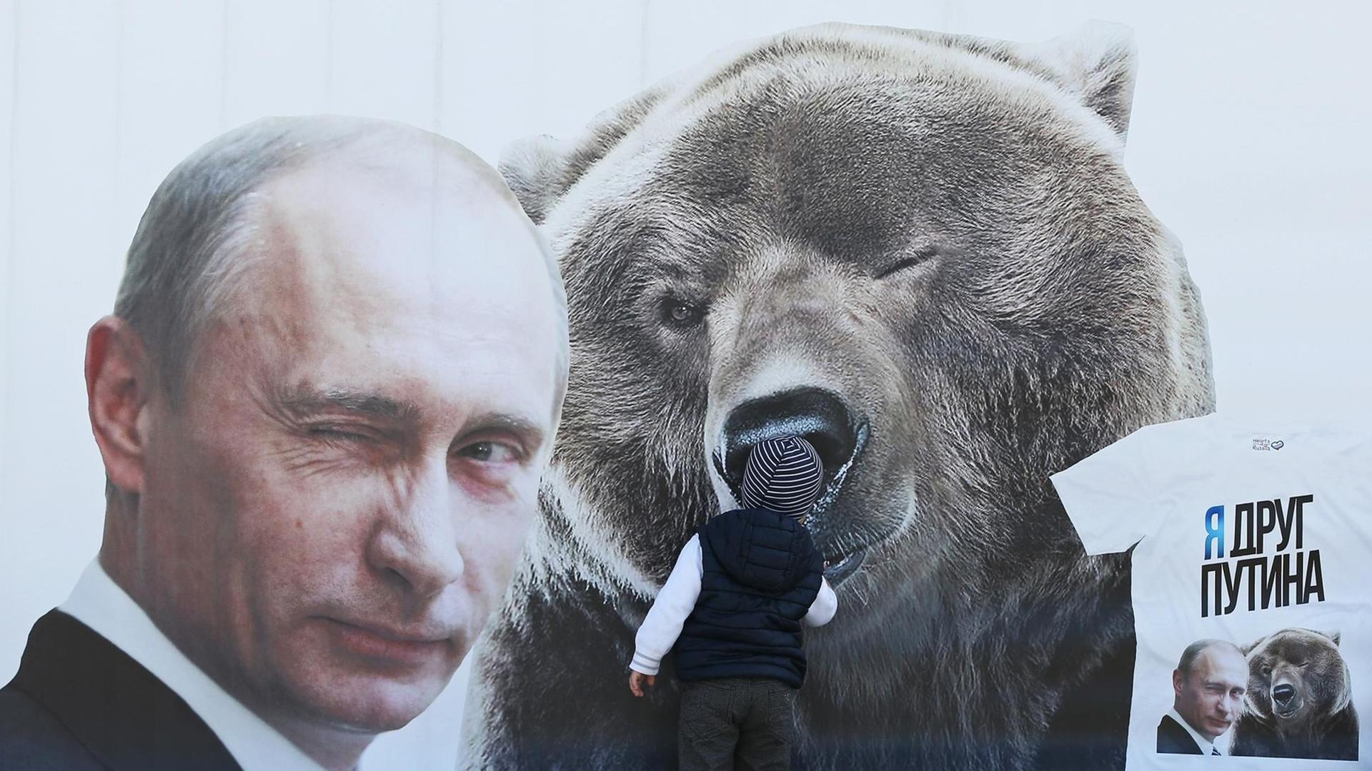Ein kleiner Junge inspiziert eine Plakatwand mit einem zwinkernden russischen Präsidenten Wladimir Putin und einem Bären, sowie einem T-Shirt mit der Aufschrift: "Ich bin ein Freund von Putin". Ein kleiner Junge inspiziert eine Plakatwand mit einem zwinkernden russischen Präsidenten Wladimir Putin und einem Bären, sowie einem T-Shirt mit der Aufschrift: "Ich bin ein Freund von Putin".