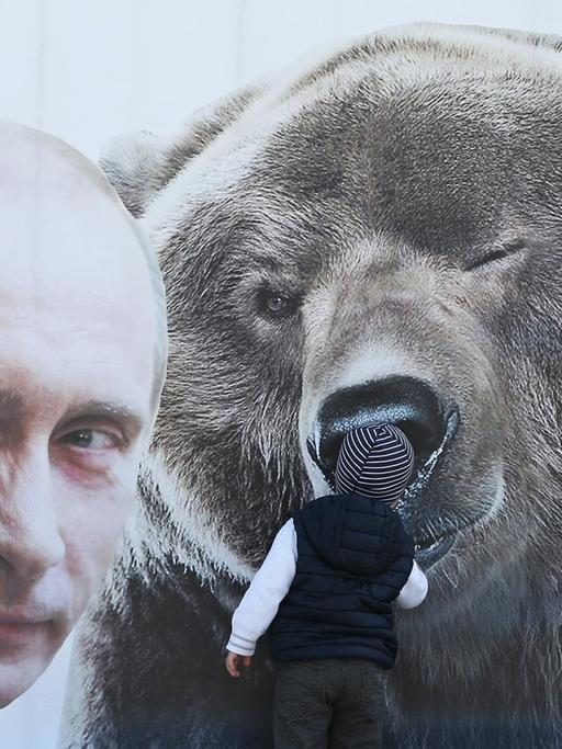 Ein kleiner Junge inspiziert eine Plakatwand mit einem zwinkernden russischen Präsidenten Wladimir Putin und einem Bären, sowie einem T-Shirt mit der Aufschrift: "Ich bin ein Freund von Putin".