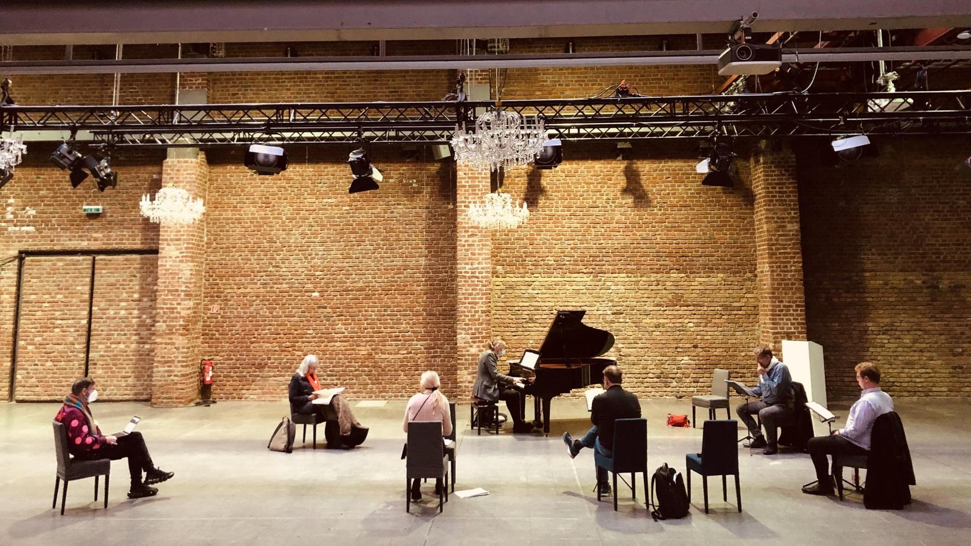 In einer Halle sitzen Menschen in einem Halbkreis um einen Flügel und haben aufgeschlagene Notenhefte vor sich. Über ihnen hängen Kronleuchter, die Wänder sind aus Backsteinen gebaut. In einer Halle sitzen Menschen in einem Halbkreis um einen Flügel und haben aufgeschlagene Notenhefte vor sich. Über ihnen hängen Kronleuchter, die Wänder sind aus Backsteinen gebaut.