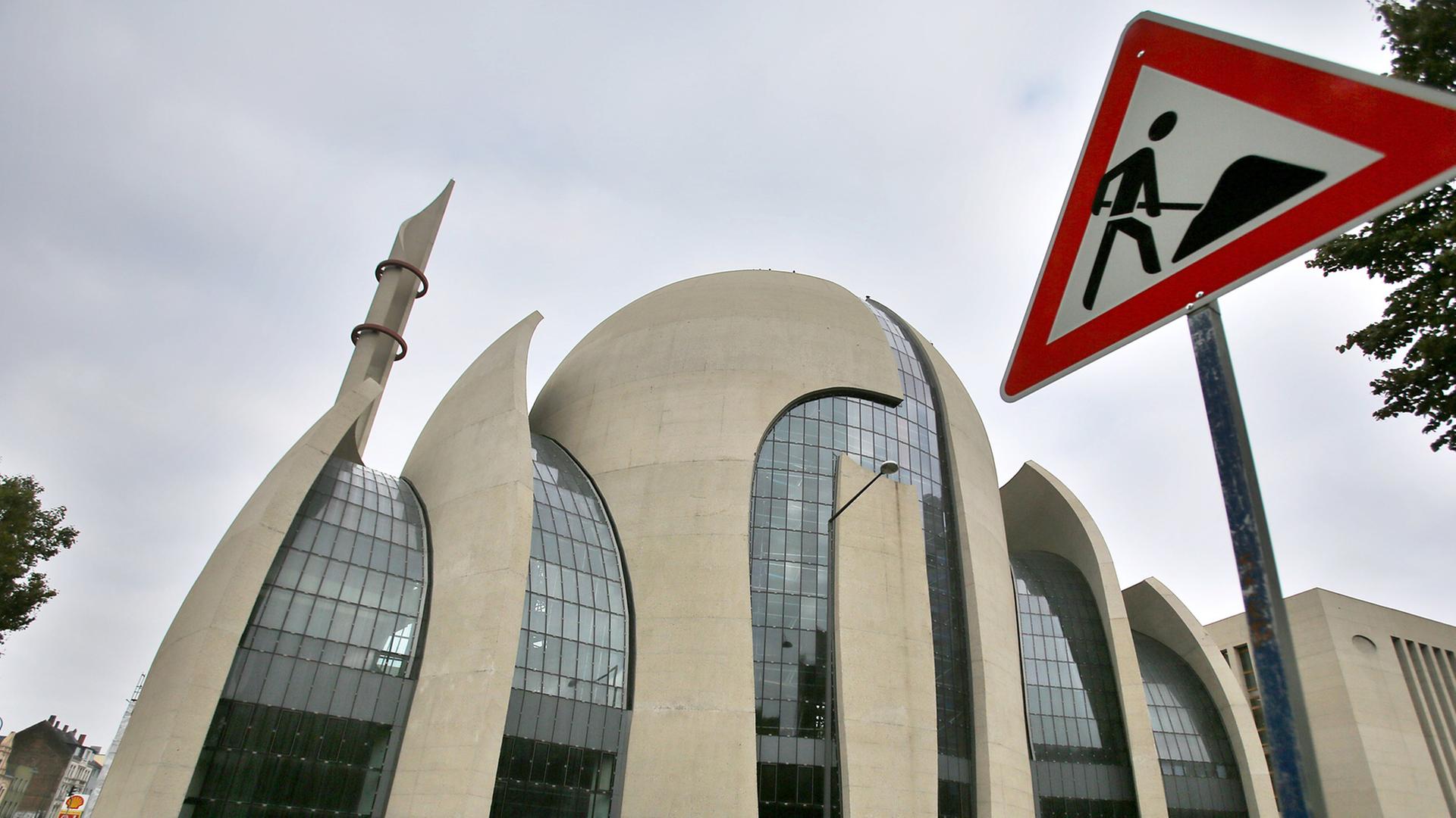 Ansicht der Kölner Zentralmoschee, eines geschwungenen Gebäudes mit Kuppel aus Beton und Glas, davor Autos auf einer Straße und ein Baustellenschild Ansicht der Kölner Zentralmoschee, eines geschwungenen Gebäudes mit Kuppel aus Beton und Glas, davor Autos auf einer Straße und ein Baustellenschild