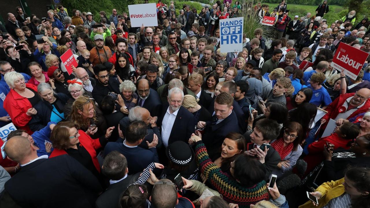 Corbyn steht inmitten einer großen Menschenmenge, um ihn herum Security-Leute. Einige Anhänger tragen "Vote Labour"-Schilder. Corbyn steht inmitten einer großen Menschenmenge, um ihn herum Security-Leute. Einige Anhänger tragen "Vote Labour"-Schilder.