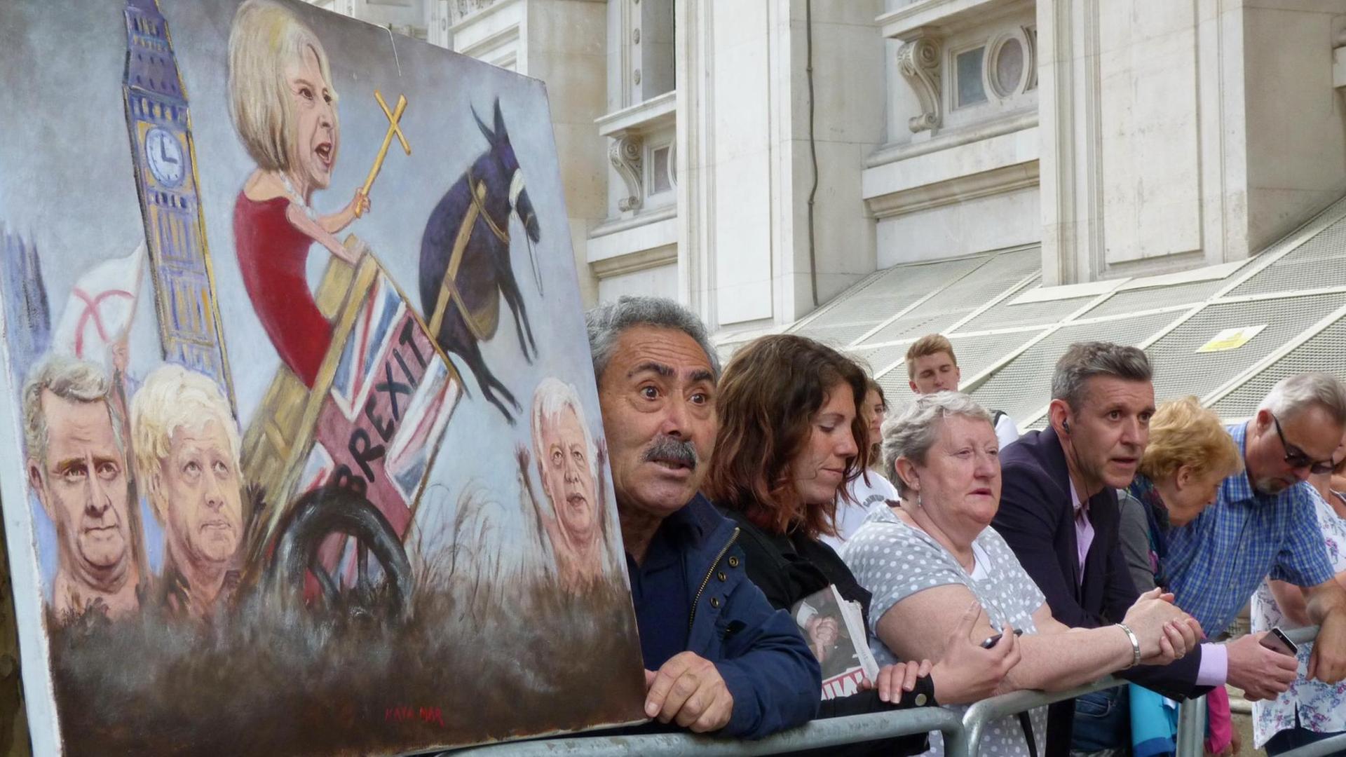 Menschen warten vor der Downing Street 10 auf Premierministerin Theresa May. Ein Mann hält eine Karrikatur von May in der Hand, die das Thema Brexit thematisiert.