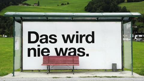 In einem leeren Bus-Wartehäuschen in einer einsamen, grünen Berglandschaft prangt ein großes Plakat mit der Aufschrift "Das wird nie was".