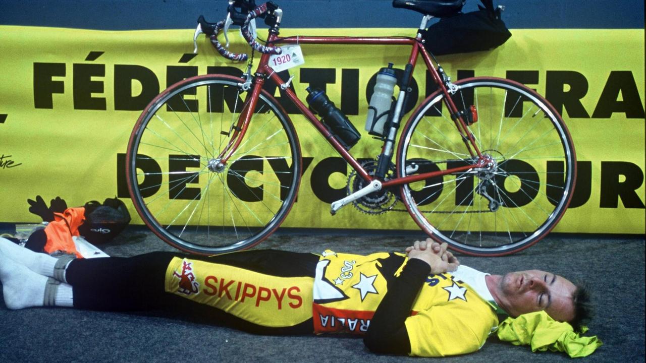 Ein Teilnehmer im gelben Trikot hat sich erschöpft auf den Boden gelegt, sein Rad lehnt neben ihm an der Wand.