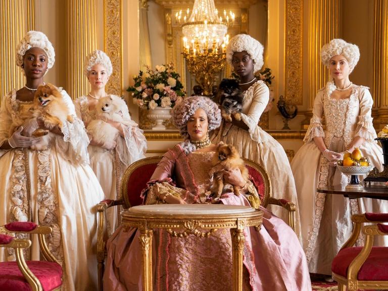 Szene aus der Serie "Bridgerton": Die Queen umringt von adligen Damen / Hofdamen, einige der Frauen sind schwarz, einige weiß.