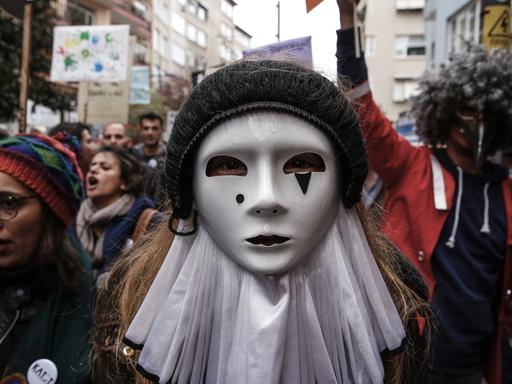 Menschen gehen demonstrierend durch eine Straße. In der Mitte des Bildes ist eine Frau oder ein Mann mit einer weißen Maske auf dem Gesicht.