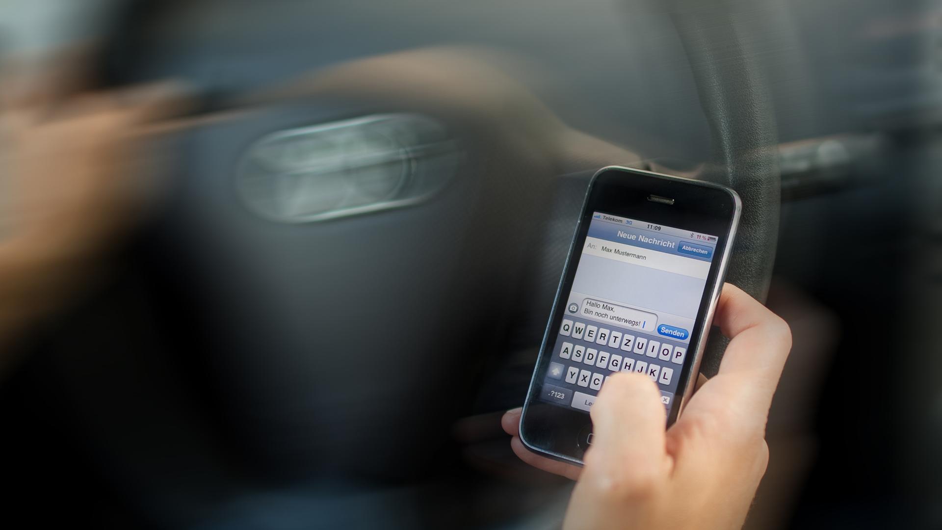 Eine Hand tippt vor einem Auto-Lenkrad in ein Handy. Eine Hand tippt vor einem Auto-Lenkrad in ein Handy.