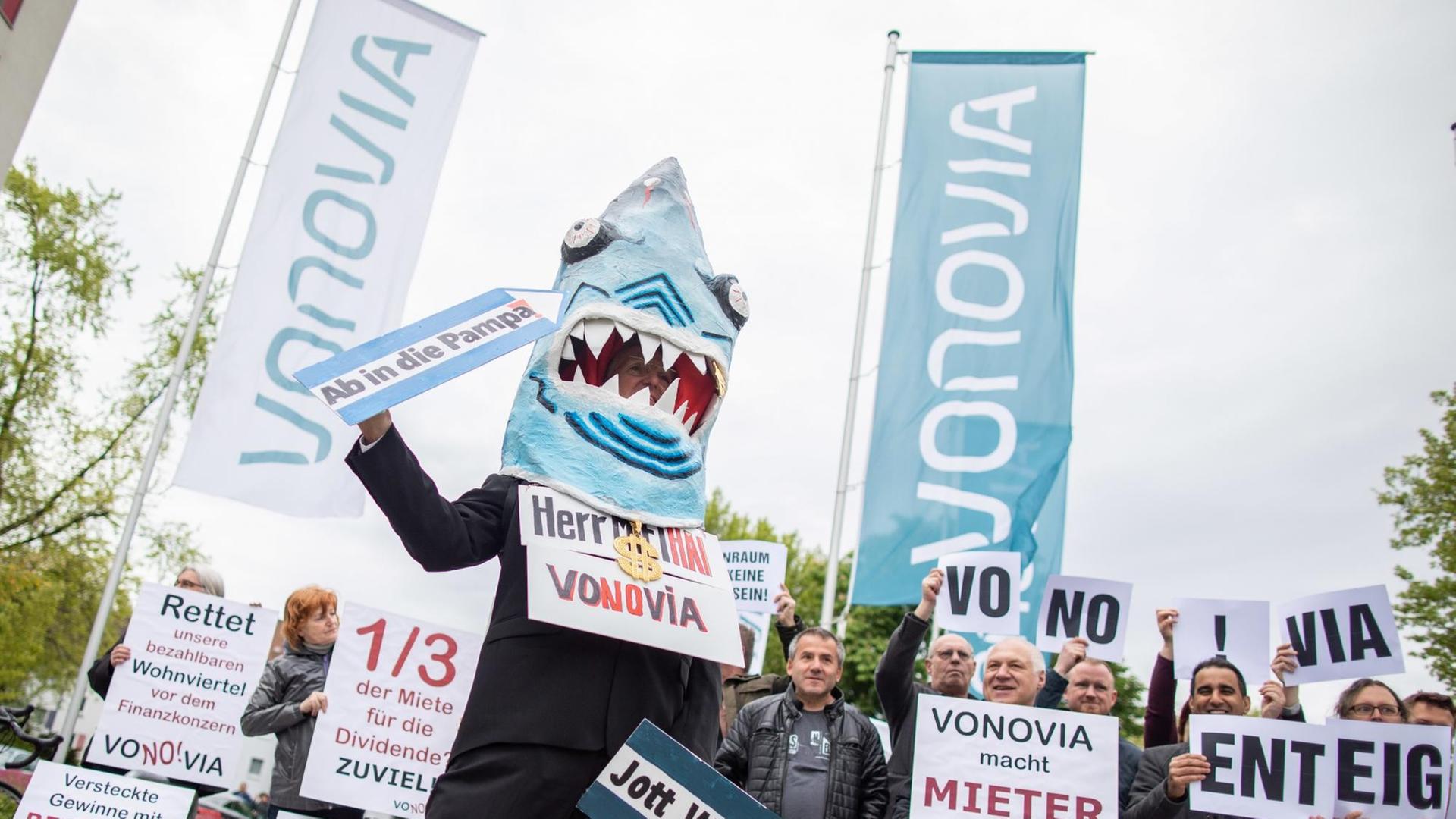 16.05.2019, Nordrhein-Westfalen, Bochum: Ein Demonstrant in einem Hai-Kostüm mit einem Schild um den Hals "Herr Miethai Vonovia" steht vor der Vonovia Hauptversammlung vor dem Ruhrcongress. Weitere Demonstranten stehen dahinter. Foto: Marcel Kusch/dpa | Verwendung weltweit 16.05.2019, Nordrhein-Westfalen, Bochum: Ein Demonstrant in einem Hai-Kostüm mit einem Schild um den Hals "Herr Miethai Vonovia" steht vor der Vonovia Hauptversammlung vor dem Ruhrcongress. Weitere Demonstranten stehen dahinter. Foto: Marcel Kusch/dpa | Verwendung weltweit