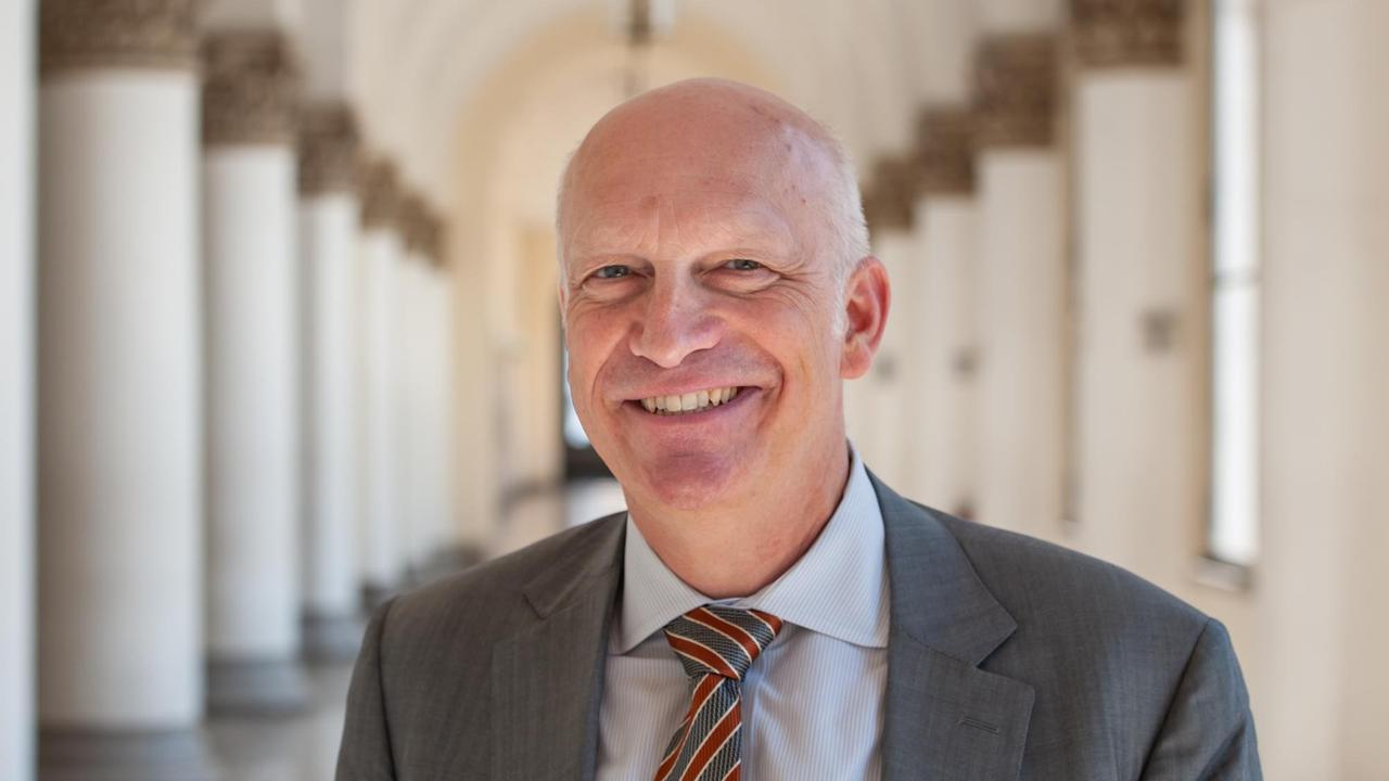 Hans van Ess, in grauem Anzug mit Krawatte, steht lächelnd in einem Säulengang. Hans van Ess, in grauem Anzug mit Krawatte, steht lächelnd in einem Säulengang.
