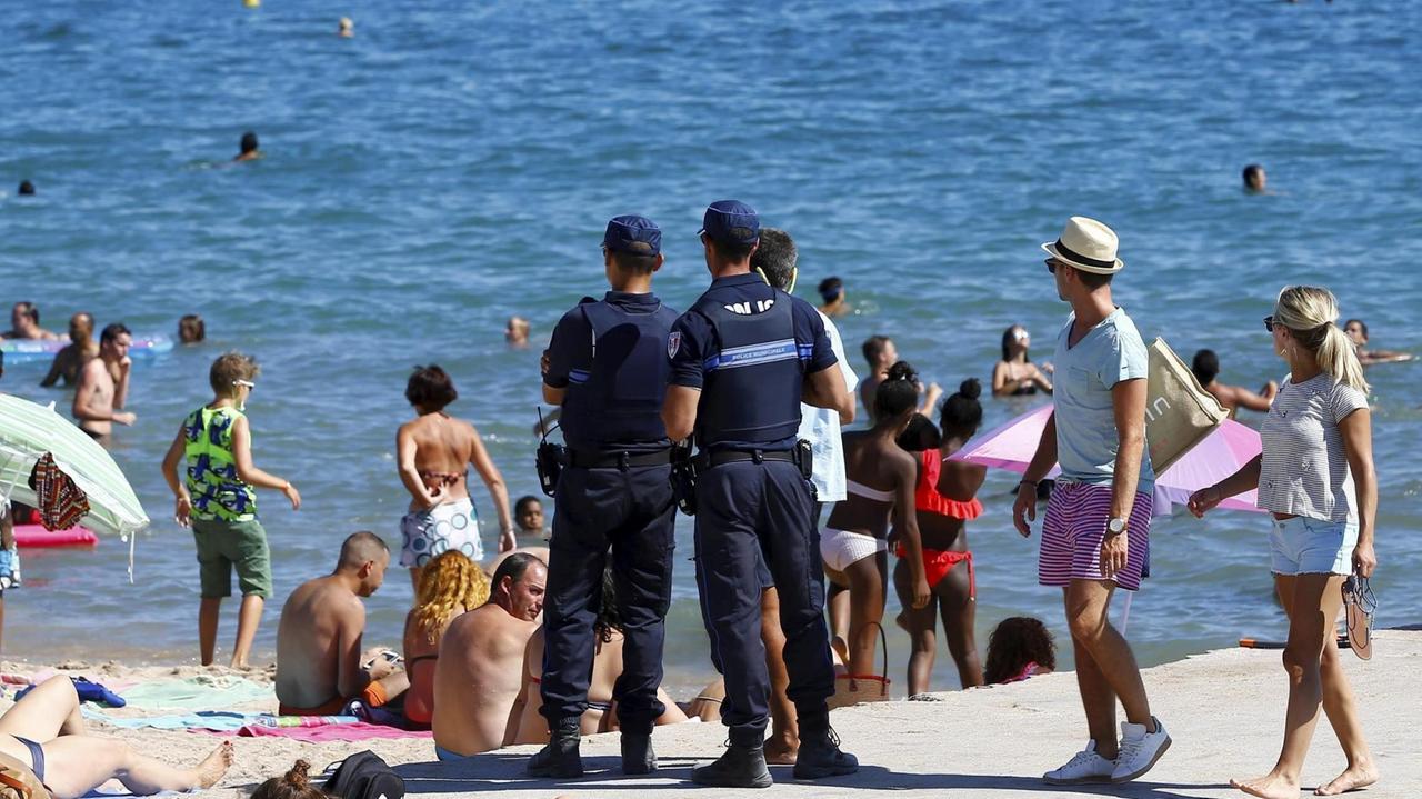 Zwei Polizisten mit blauer Uniform stehen mit Rücken zur kamera am Strand von Cannes. Im Wasser sind Badende zu sehen, am Strand Urlauber in sommerlicher Kleidung.