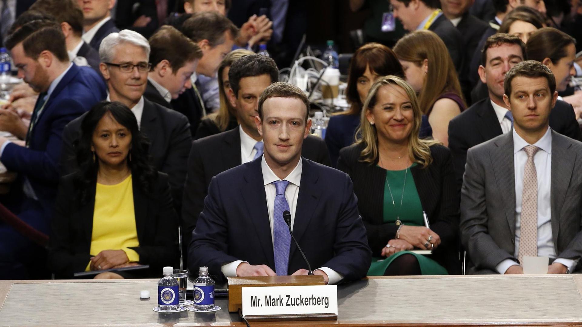 Mark Zuckerberg sitzt an einem Tisch in einem Raum des US-Kongresses, hinter ihm eine Gruppe von Menschen Mark Zuckerberg sitzt an einem Tisch in einem Raum des US-Kongresses, hinter ihm eine Gruppe von Menschen