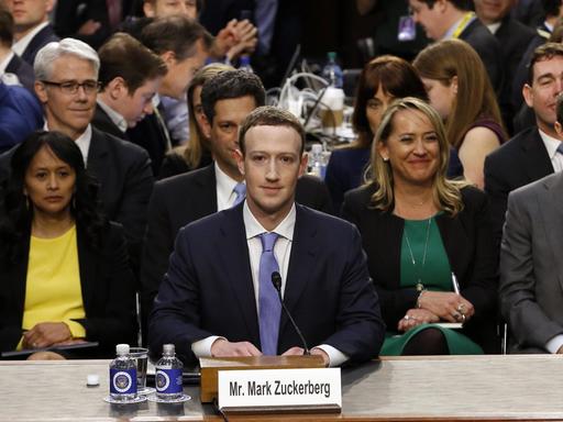 Mark Zuckerberg sitzt an einem Tisch in einem Raum des US-Kongresses, hinter ihm eine Gruppe von Menschen