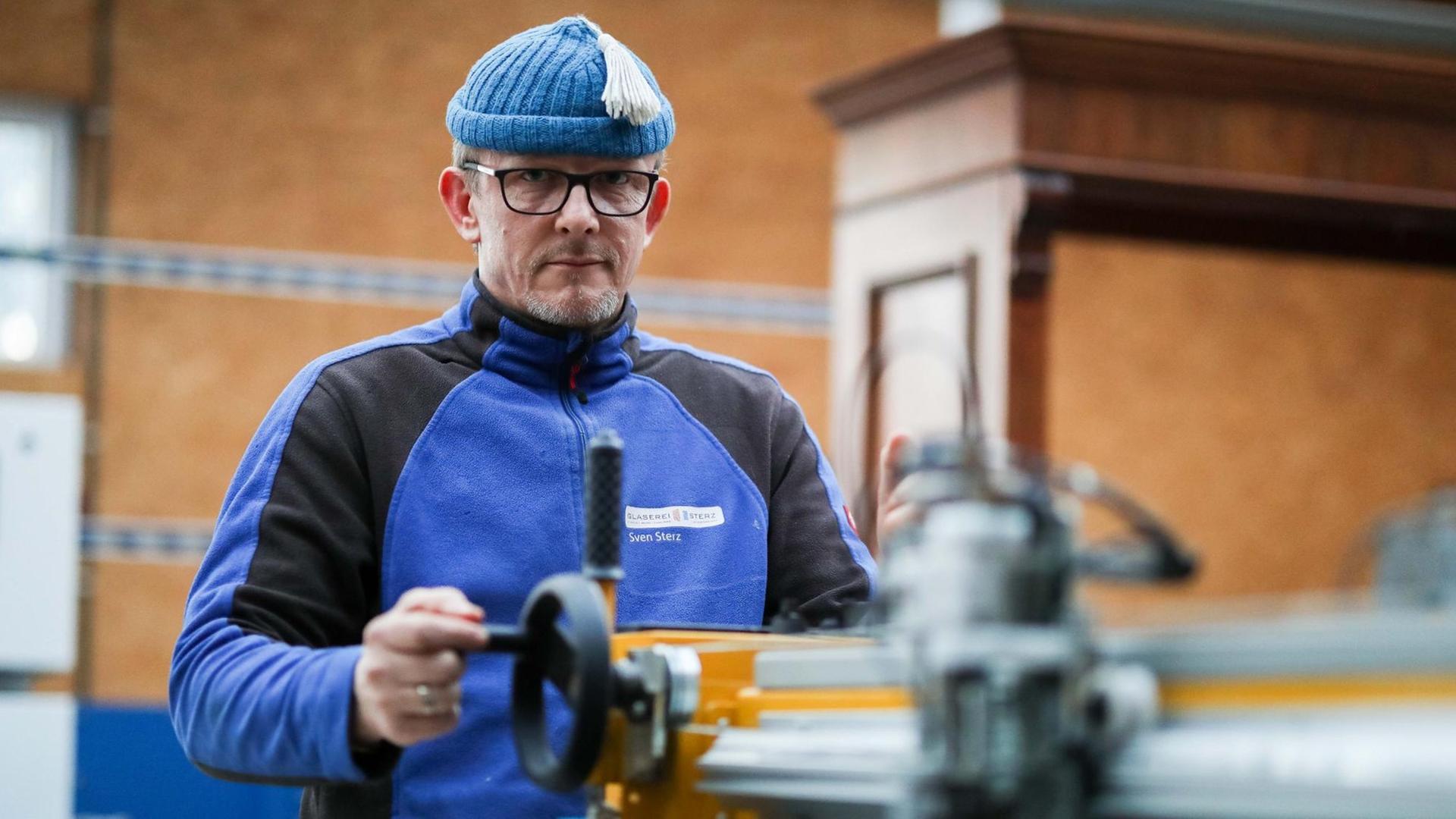 Glasermeister Sven Sterz steht in seiner Werkstatt an einem Glasschneidetisch und wirft einen Blick in die Kamera. Glasermeister Sven Sterz steht in seiner Werkstatt an einem Glasschneidetisch und wirft einen Blick in die Kamera.