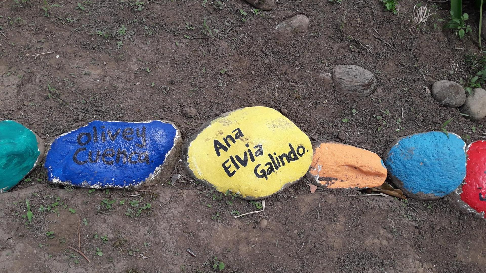 Kieselsteine, blau, gelb und in weiteren Farben bemalt, tragen die Namen von kolumbianischen Landbewohnern, die ermordet wurden. Sie liegen in einer Kette auf dem staubigen Erdboden. Kieselsteine, blau, gelb und in weiteren Farben bemalt, tragen die Namen von kolumbianischen Landbewohnern, die ermordet wurden. Sie liegen in einer Kette auf dem staubigen Erdboden.