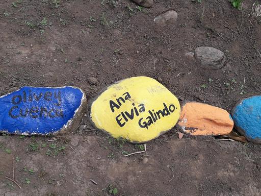 Kieselsteine, blau, gelb und in weiteren Farben bemalt, tragen die Namen von kolumbianischen Landbewohnern, die ermordet wurden. Sie liegen in einer Kette auf dem staubigen Erdboden.
