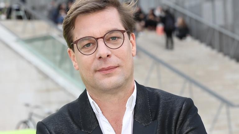 Alexander Brüggemann steht vor einem Gebäude der Kunst Stiftung Ludwig Wien. Er trägt Brille und hat helles, kurzes, gescheiteltes Haar. Er hat ein weißes Hemd an, das nicht zugeknöpft ist, und trägt dazu einen Smoking.