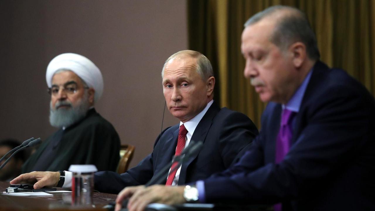 Die drei sitzen nebeneinander an Mikrofonen. Putin in der Mitte sieht man scharf, die beiden anderen an den Seiten unscharf. Erdogan scheint zu reden. Die drei sitzen nebeneinander an Mikrofonen. Putin in der Mitte sieht man scharf, die beiden anderen an den Seiten unscharf. Erdogan scheint zu reden.