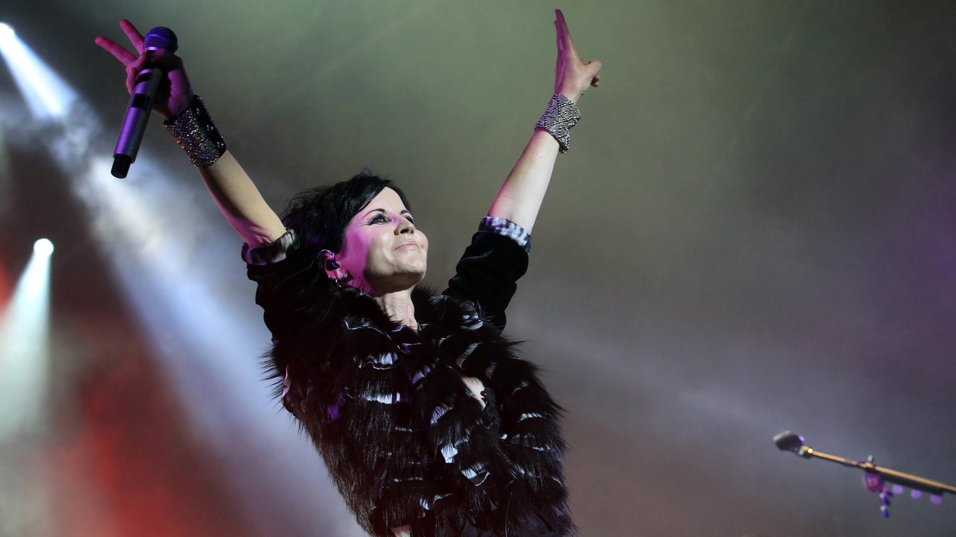 Dolores O’Riordan steht mit nach oben gereckten Armen auf der Bühne, in einer Hand hält sie ein Mikrofon. Dolores O’Riordan steht mit nach oben gereckten Armen auf der Bühne, in einer Hand hält sie ein Mikrofon.