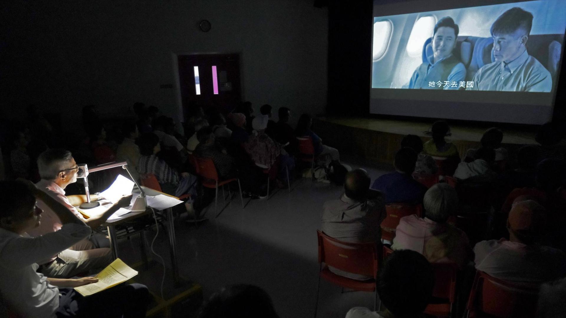 Ein Mann an einem Pult mit Mikrofon beschreibt die Handlung eines auf einer Leinwand abgespielten Films, vor ihm in mehreren Stuhlreihen das blinden bzw. sehbehinderte Publikum Ein Mann an einem Pult mit Mikrofon beschreibt die Handlung eines auf einer Leinwand abgespielten Films, vor ihm in mehreren Stuhlreihen das blinden bzw. sehbehinderte Publikum