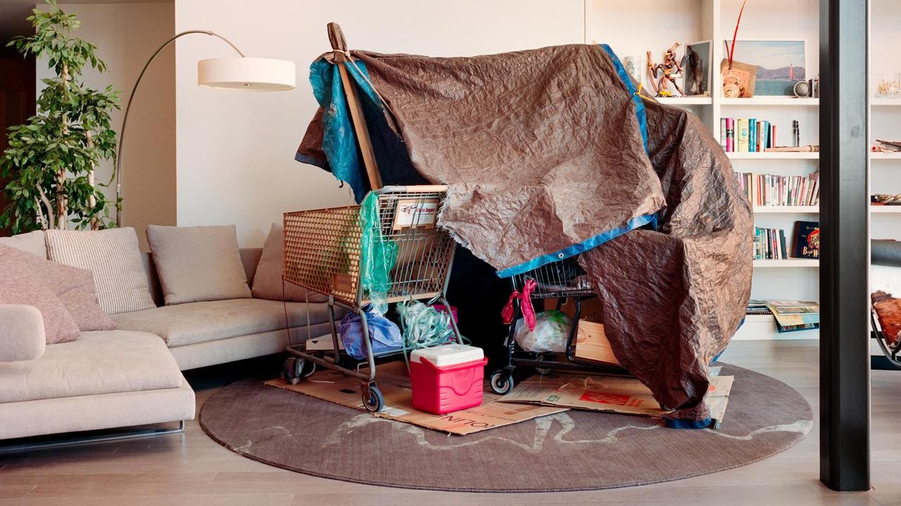 Aus Holzstöcken, einem Einkaufwagen und einer zerschlissenen Plane ist ein Zelt gebaut worden, das in einem eleganten Wohnzimmer mit beigem Teppich und farblich passende Sofa steht. Aus Holzstöcken, einem Einkaufwagen und einer zerschlissenen Plane ist ein Zelt gebaut worden, das in einem eleganten Wohnzimmer mit beigem Teppich und farblich passende Sofa steht.