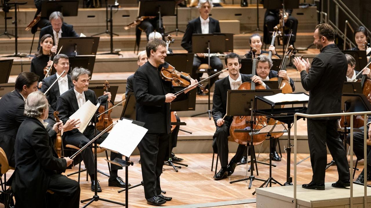 Ein Geiger steht mit seinem Instrument im Umfeld des Orchesters und erhält Beifall von Kollegen und dem Dirigenten.