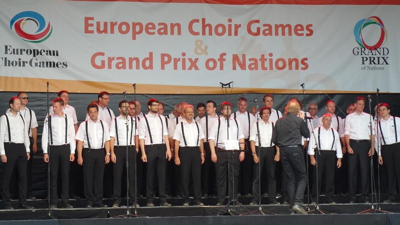 Die Männer des Chores stehen bei den Choir Games in schwarzen Hosen und weißen Hemden auf der Bühne. Die Männer des Chores stehen bei den Choir Games in schwarzen Hosen und weißen Hemden auf der Bühne.
