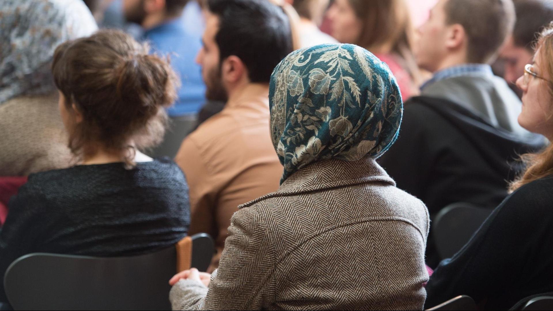 Das Bild zeigt eine Frau mit Kopftuch, die zwischen anderen Menschen im Publikum einer Veranstaltung sitzt.