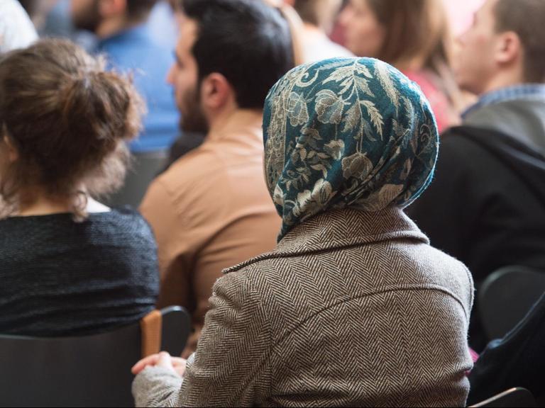 Das Bild zeigt eine Frau mit Kopftuch, die zwischen anderen Menschen im Publikum einer Veranstaltung sitzt.