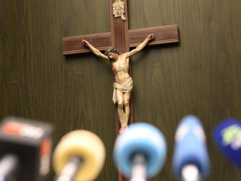 Ein Kreuz hängt bei einer Pressekonferenz vor Mikrofonen an einer Wand.