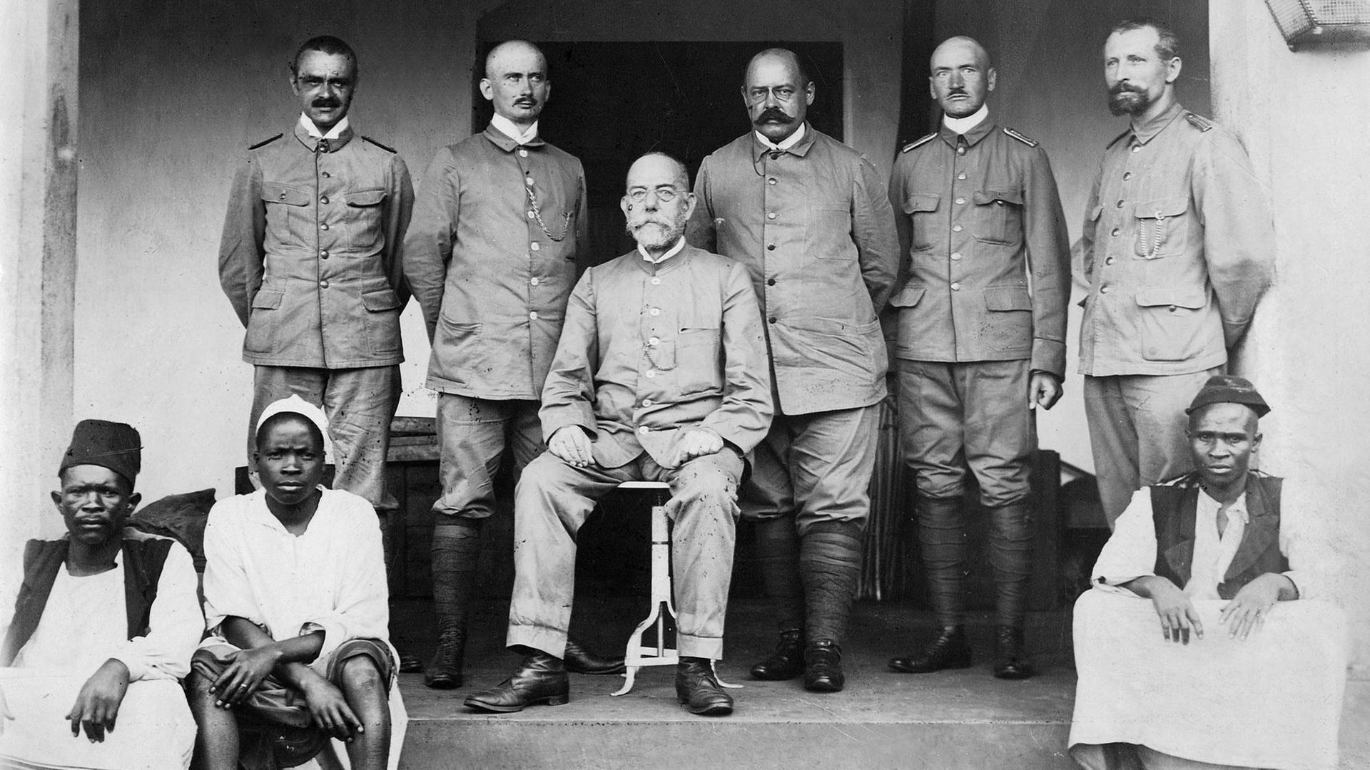 Robert Koch (sitzend) mit Mitgliedern der Expedition zur Untersuchung der Schlafkrankheit in Afrika; v.l.: Dr. Kudicke, Prof. Klein, Koch, Prof. Beck, Stabsarzt Pause, Feldwebel Sacher (Foto: Franz-Otto-Koch - 1897) Vor den Weißen hocken drei Schwarze auf den Stufen der Terrasse. Robert Koch (sitzend) mit Mitgliedern der Expedition zur Untersuchung der Schlafkrankheit in Afrika; v.l.: Dr. Kudicke, Prof. Klein, Koch, Prof. Beck, Stabsarzt Pause, Feldwebel Sacher (Foto: Franz-Otto-Koch - 1897) Vor den Weißen hocken drei Schwarze auf den Stufen der Terrasse.