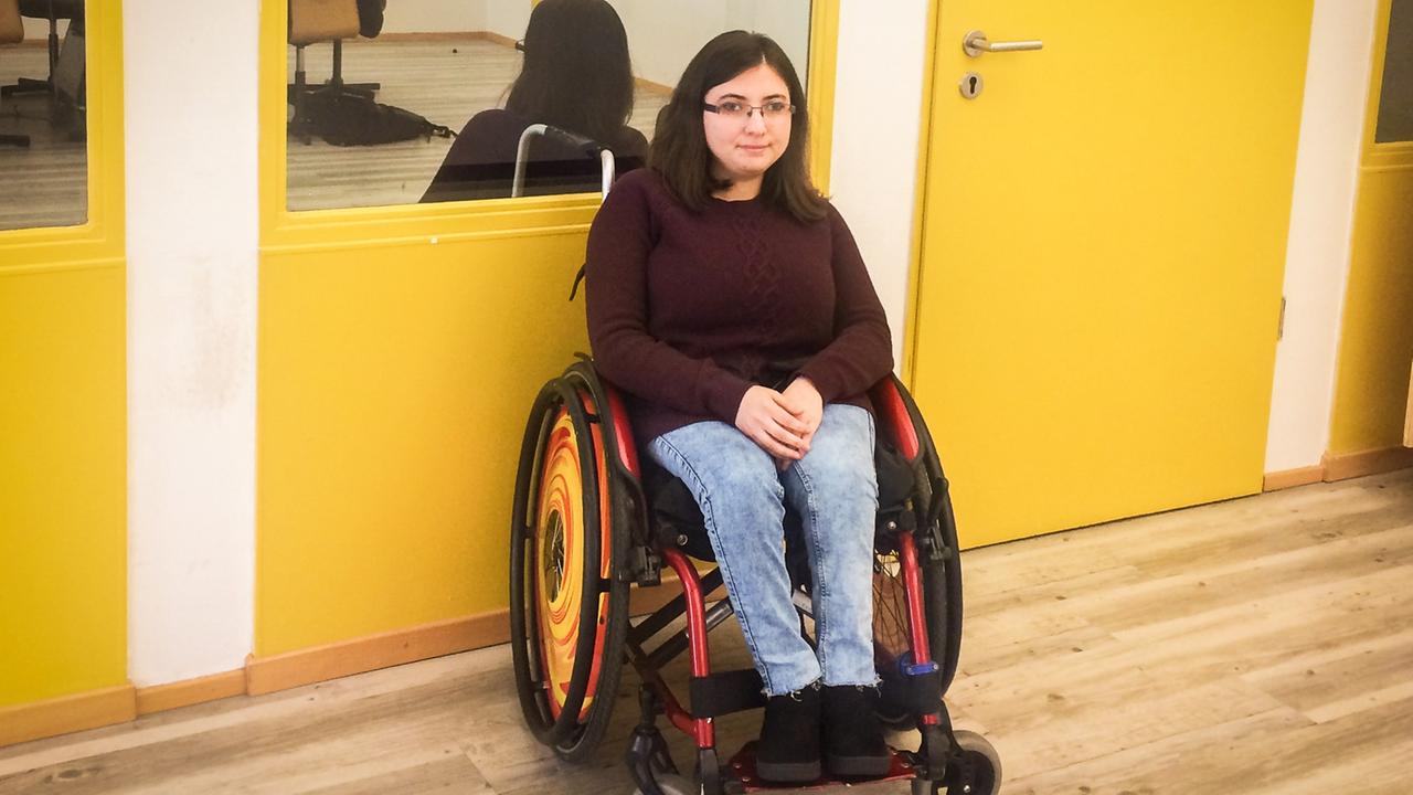 Duygu Oezen sitzt in Ihrem Rollstuhl vor einer gelb gestrichenen Wand. Duygu Oezen sitzt in Ihrem Rollstuhl vor einer gelb gestrichenen Wand.