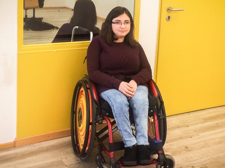 Duygu Oezen sitzt in Ihrem Rollstuhl vor einer gelb gestrichenen Wand.