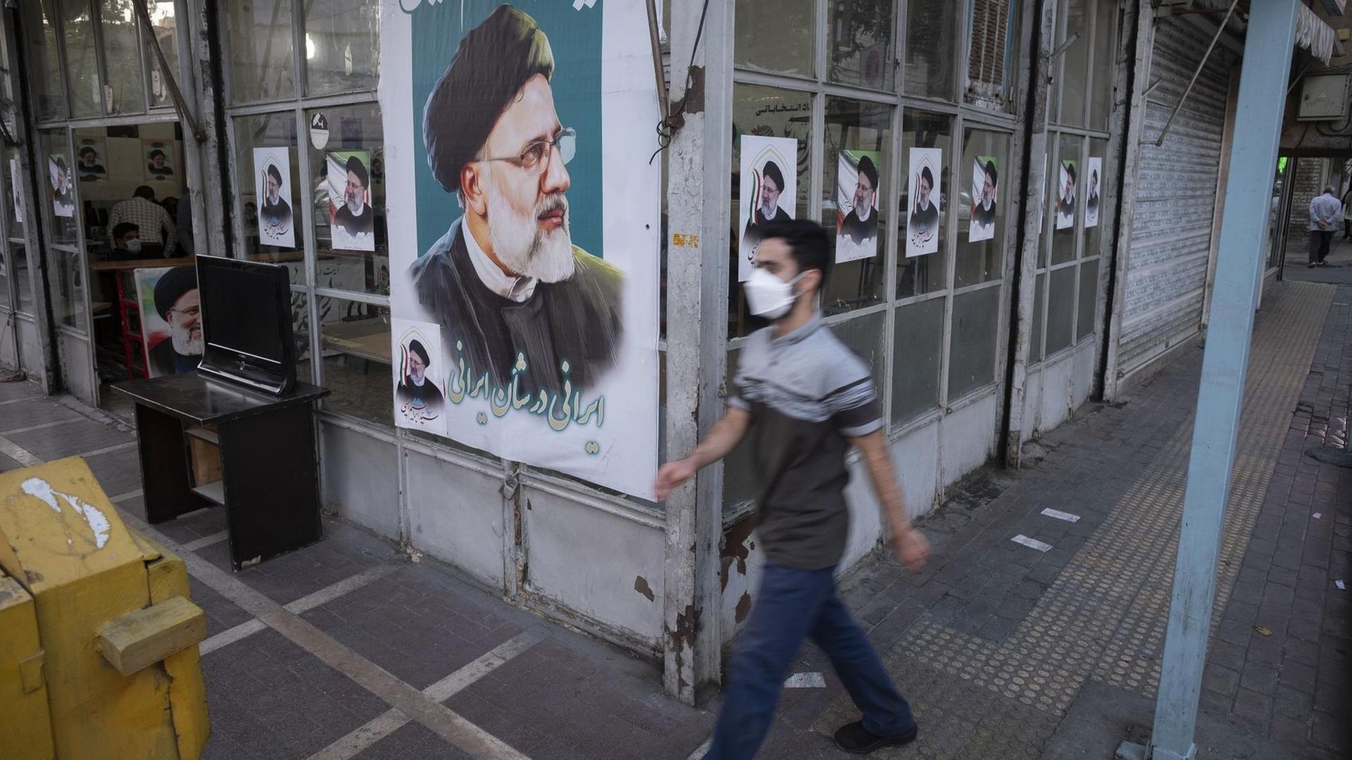 Ein Mann läuft an der Fassade eines Geschäfts vorbei, die mit Wahlplakaten von Ebrahim Raisi beklebt ist. Ein Mann läuft an der Fassade eines Geschäfts vorbei, die mit Wahlplakaten von Ebrahim Raisi beklebt ist.