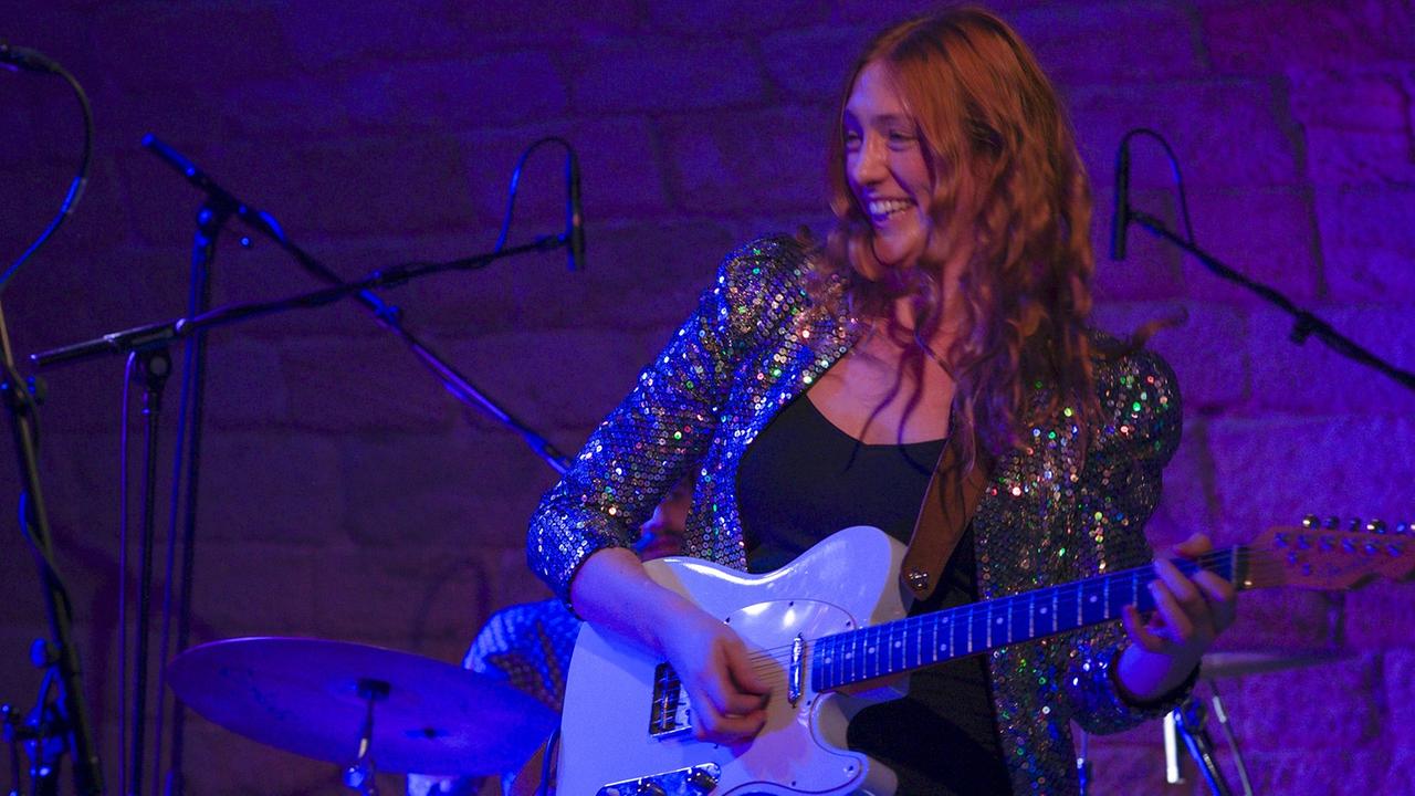 Ein Frau mit langen roten Locken und pailettenbesetztem Jackett steht lächelnd auf einer Bühne und spielt eine weiße E-Gitarre. Ein Frau mit langen roten Locken und pailettenbesetztem Jackett steht lächelnd auf einer Bühne und spielt eine weiße E-Gitarre.