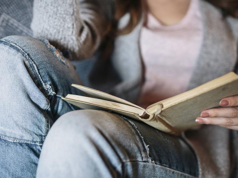 Eine Frau sitzt lesend auf einem Sofa. Das Buch ist der Mittelpunkt des Bildes.