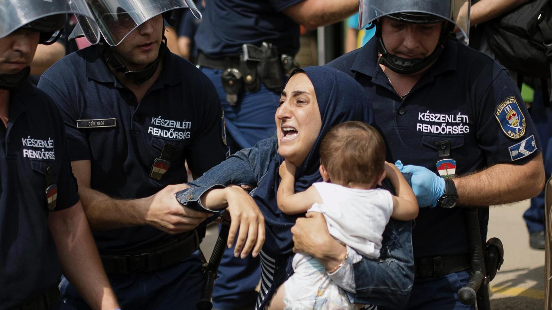 Polizisten in Schutzkleidung und Helm zerren an einer muslimischen Frau mit ihrem Kleinkind auf dem Arm. Polizisten in Schutzkleidung und Helm zerren an einer muslimischen Frau mit ihrem Kleinkind auf dem Arm.