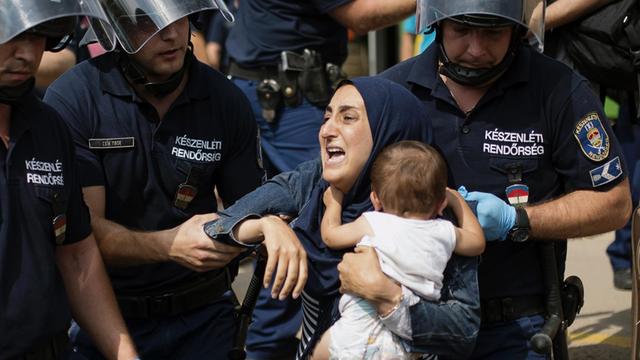 Polizisten in Schutzkleidung und Helm zerren an einer muslimischen Frau mit ihrem Kleinkind auf dem Arm.