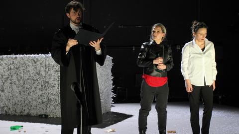 Fabian Eyer, Nadine Geyersbach und Irene Kleinschmidt (v.l.) im "Aus dem Nichts" am Theater Bremen in der Regie von Nurkan Erpulat. Ein Schauspieler und zwei Schauspielerinnen stehen auf einer Bühne. Der Mann spricht in ein Mikrofon.