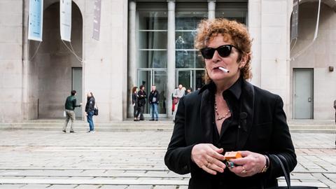 Die Künstlerin Nan Goldin steht vor einem Museum und raucht eine Zigarette.